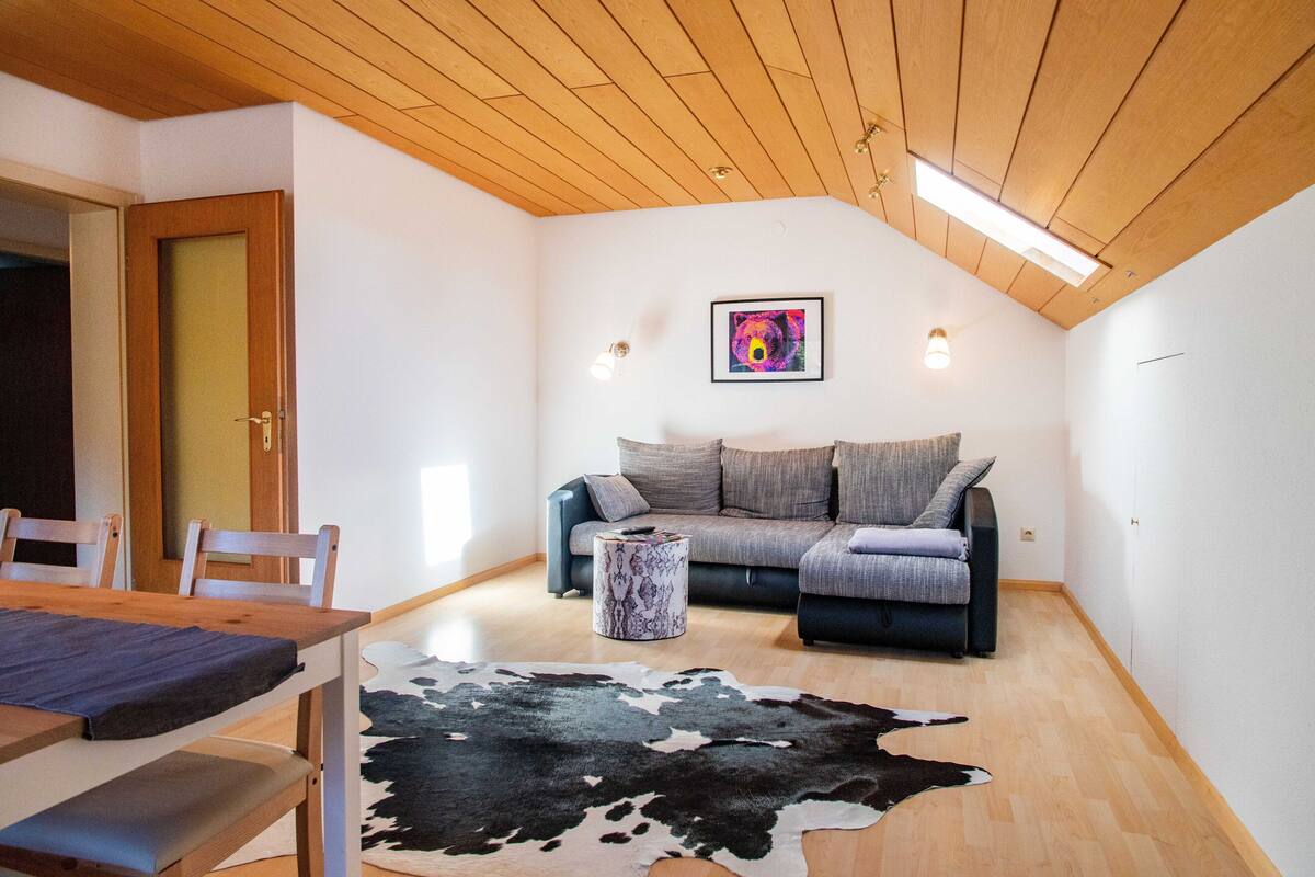 Een gezellige woonkamer met een houten plafond, een bank, een gestreept voetenbankje en een koeienhuid vloerkleed.