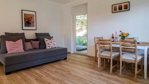 Un salon moderne avec un canapé gris, une table à manger en bois et des chaises. Un tableau encadré est accroché au mur, et une porte donne accès à un jardin.