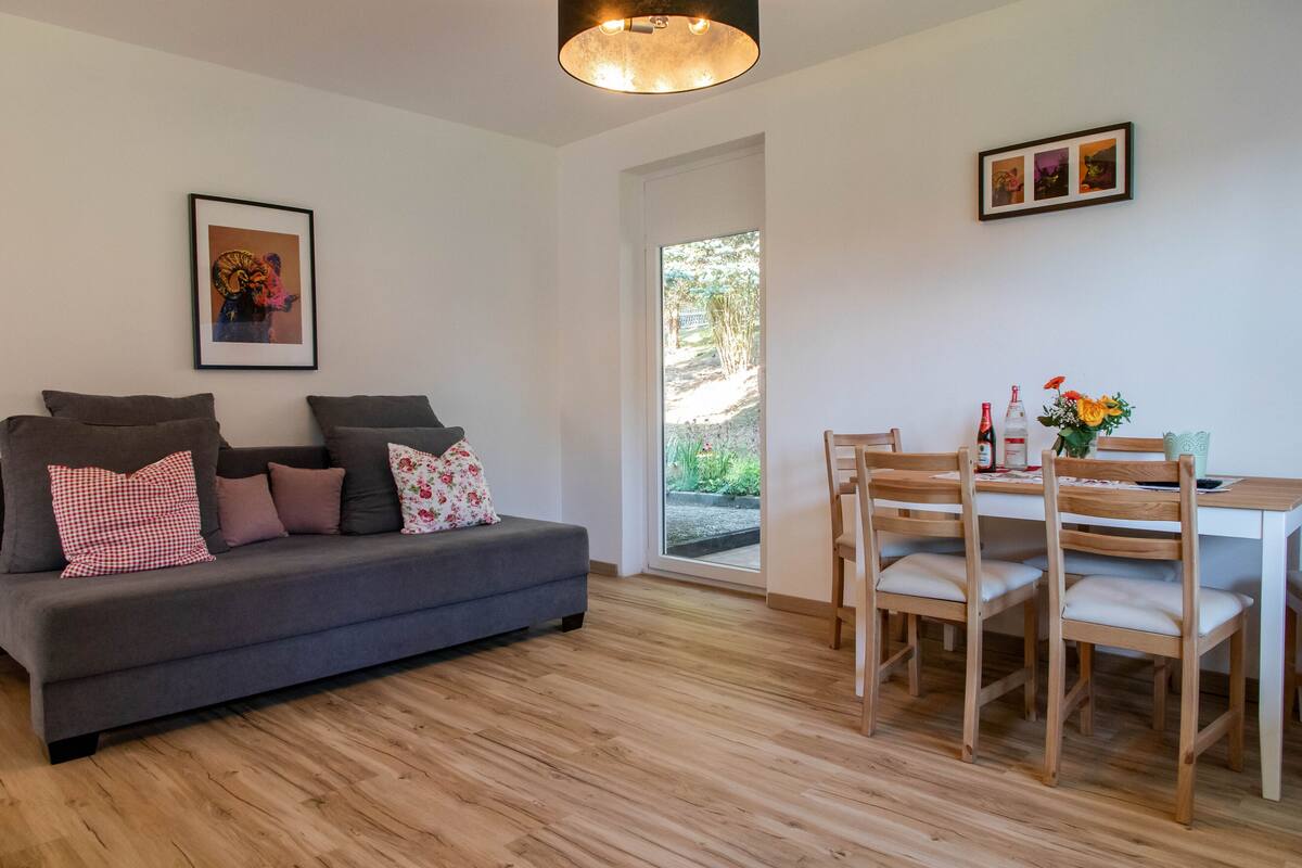 Een moderne woonkamer met een grijze bank, een houten eettafel en stoelen. Er hangt een lijst met een foto aan de muur en een deur die naar een tuin leidt.