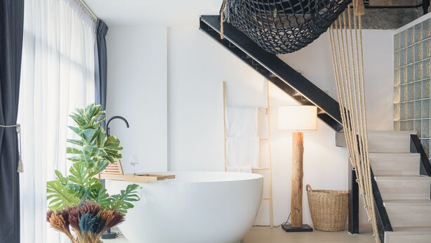 Un baño moderno con una bañera grande, un tocador de madera, una planta en maceta y una escalera con barandilla de cuerda.