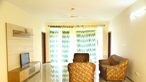 A spacious living room with a flat-screen TV on a wooden cabinet, two patterned armchairs, and a ceiling fan. - TripThrill Maytas Hill Apartments (Hyderabad)