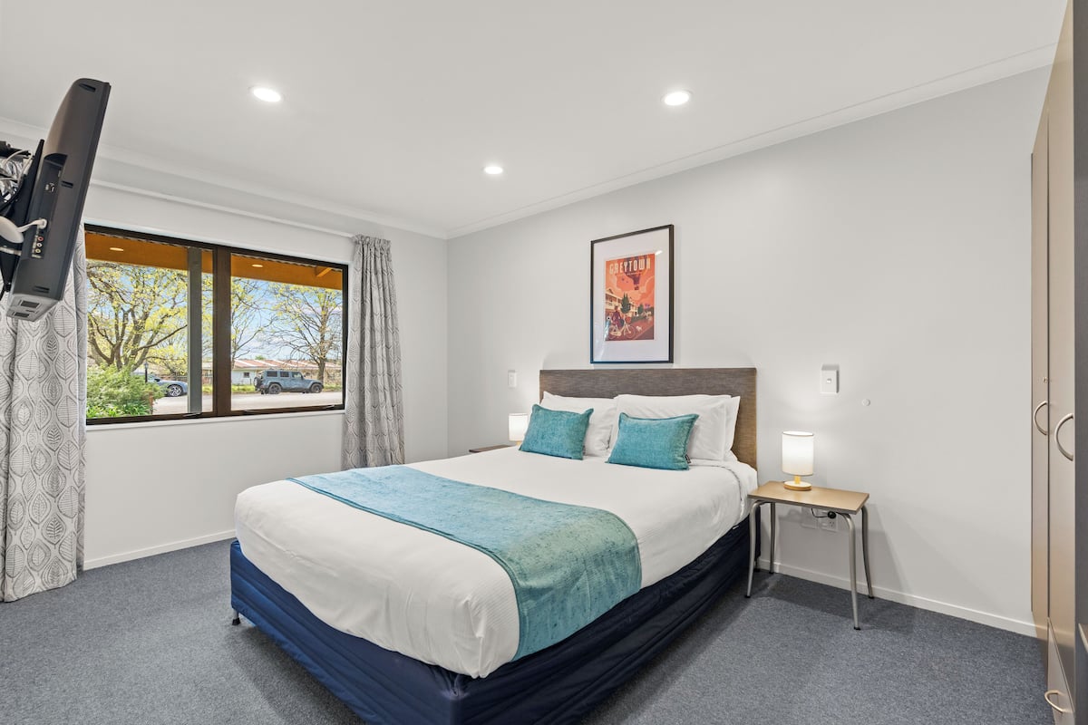 A hotel room with a large bed, a bedside table with a lamp, a window with curtains, and a framed picture on the wall.