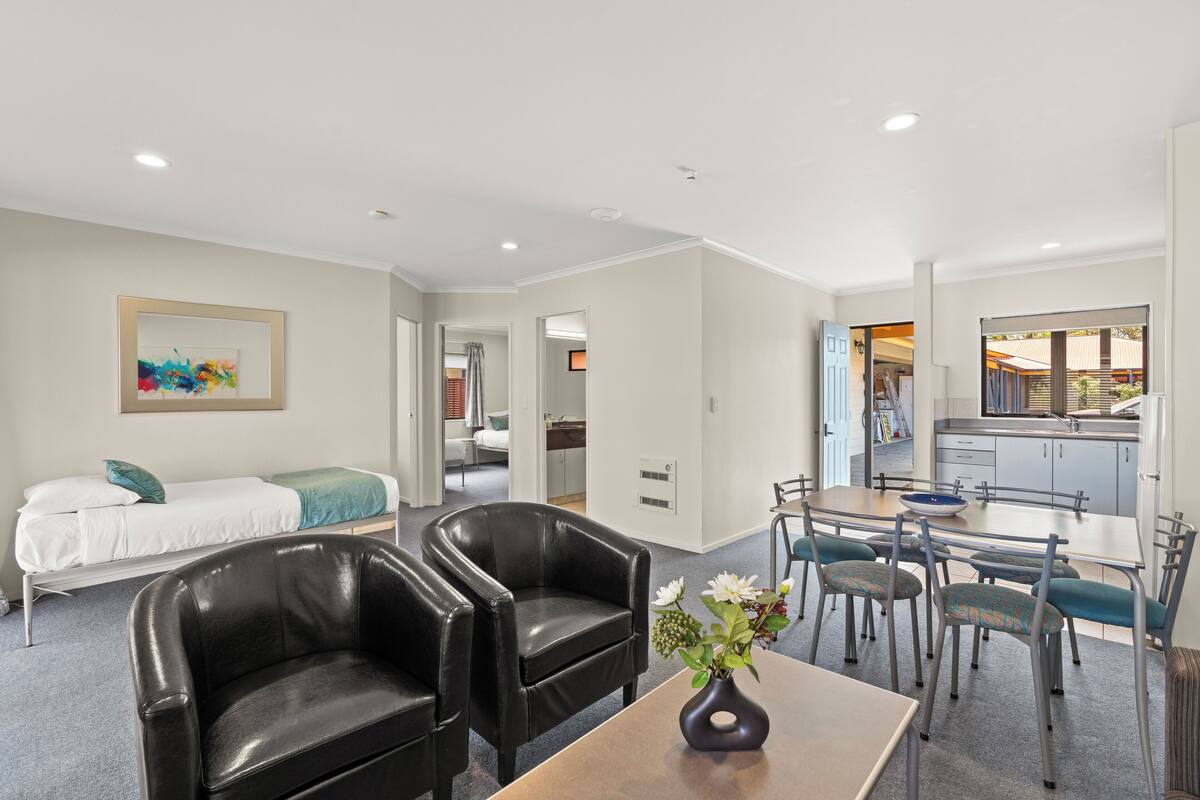 A modern hotel room with a bed, a dining area with a table and chairs, a kitchen area, and a living room with a sofa and a painting on the wall.