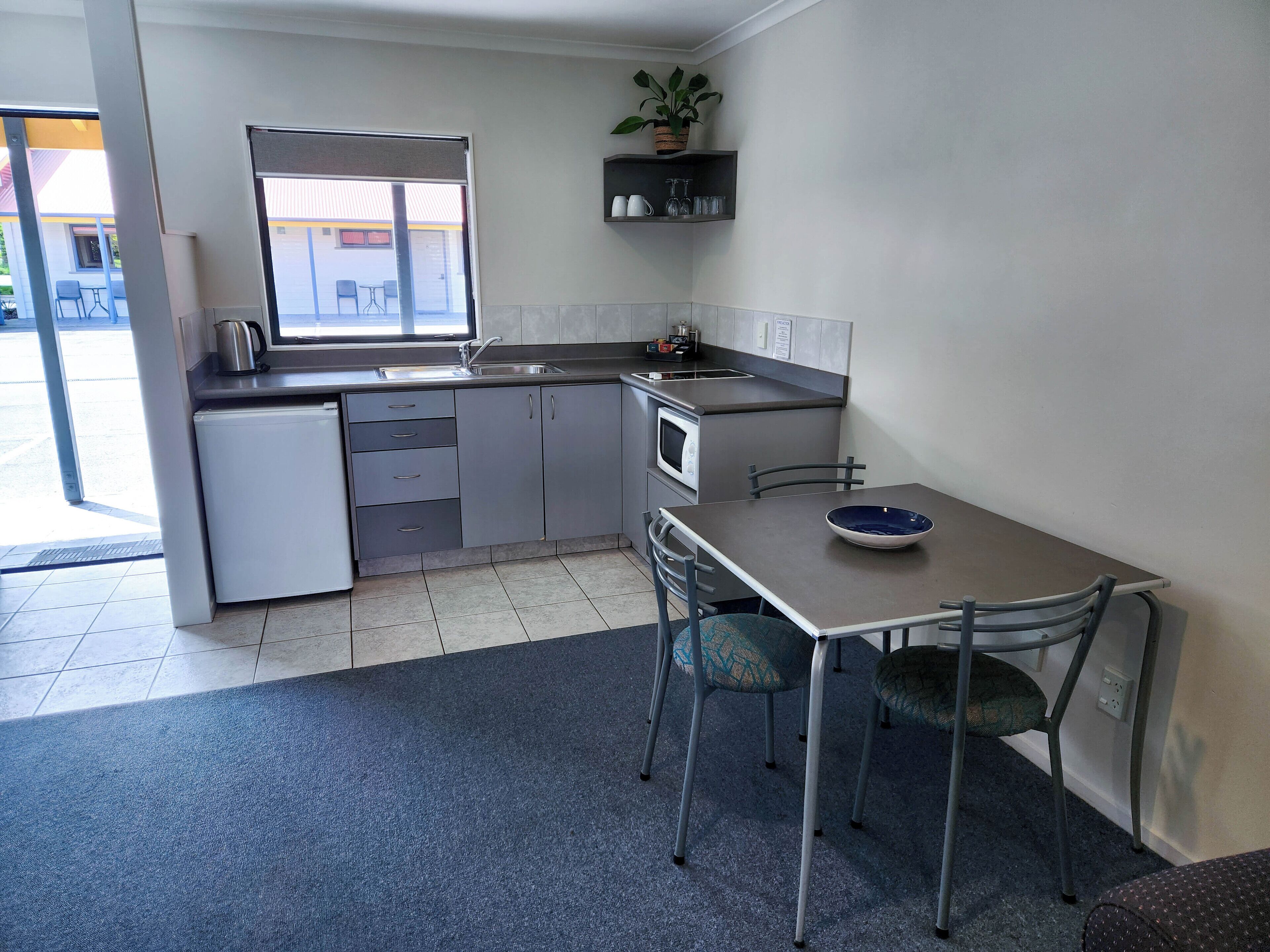 Cocina compacta con lavamanos, microondas y un refrigerador pequeño. Hay una mesa de comedor con sillas y una planta en maceta sobre una repisa.