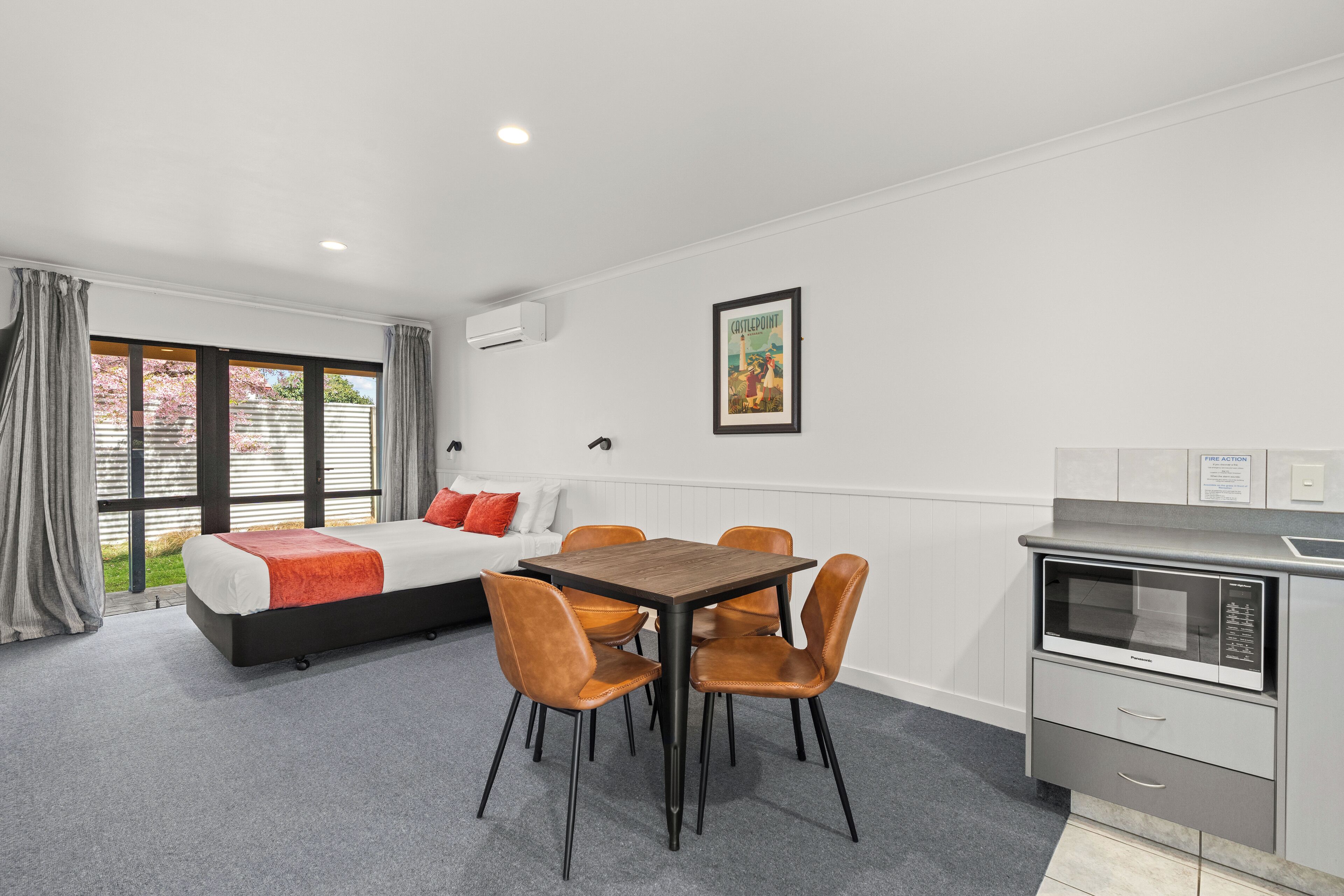 Una habitación moderna con cama, una mesa de comedor con sillas, una kitchenette y un amplio ventanal con cortinas.