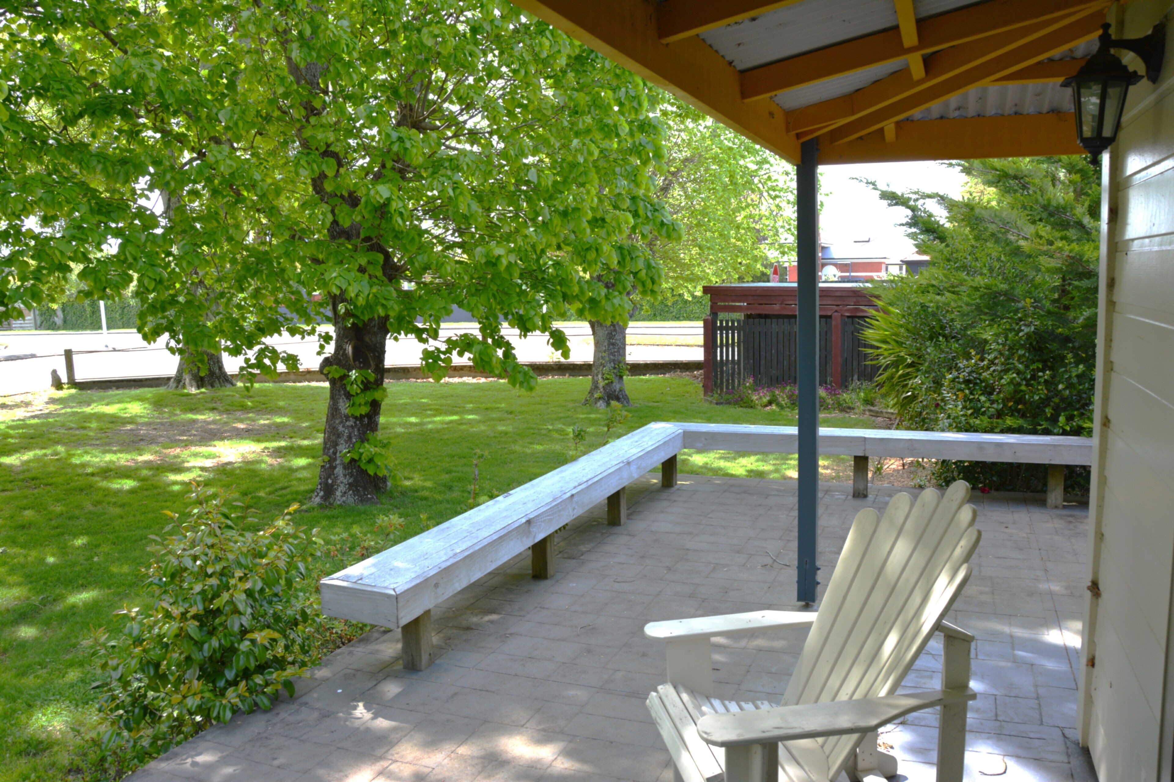 An outdoor patio with a wooden bench and a white Adirondack chair, surrounded by greenery and a building in the background.