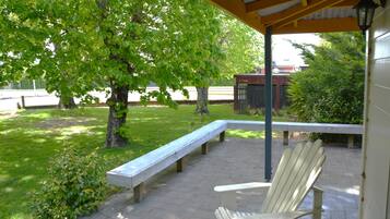An outdoor patio with a wooden bench and a white Adirondack chair, surrounded by greenery and a building in the background.