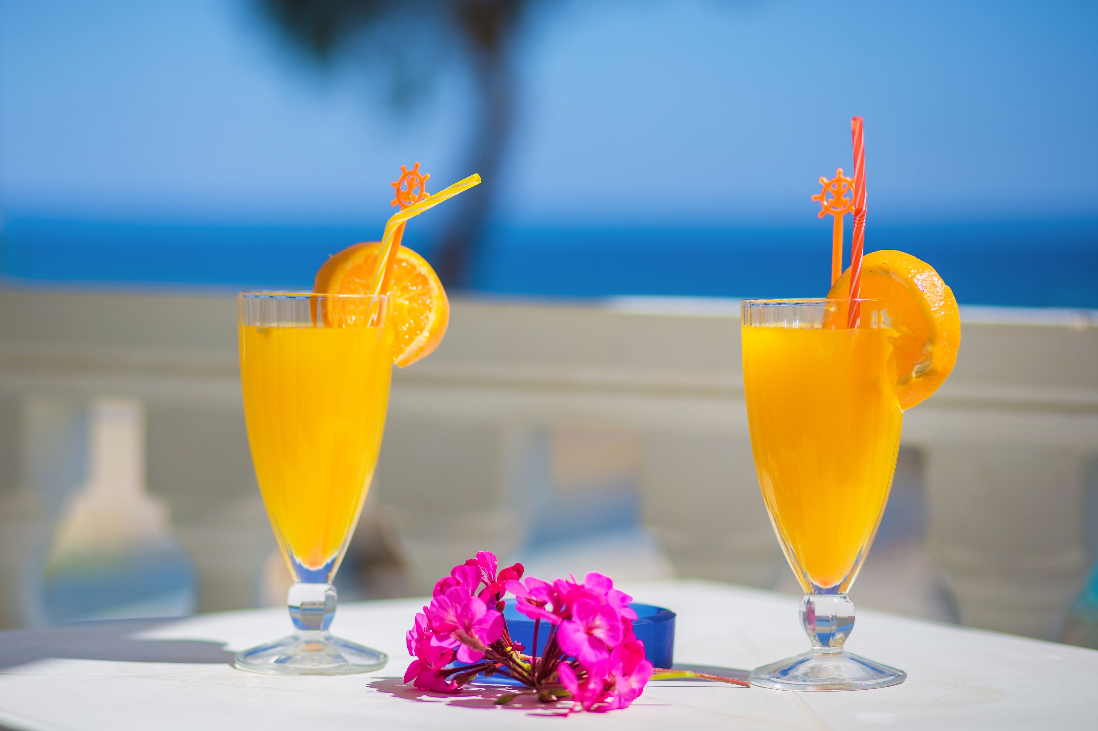 Two tall glasses of orange juice with orange slices and straws, placed on a table with a view of the ocean.