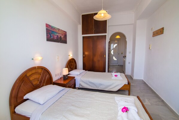 A hotel room with two single beds, wooden headboards, a wooden wardrobe, a painting on the wall, and a hallway leading to another room.