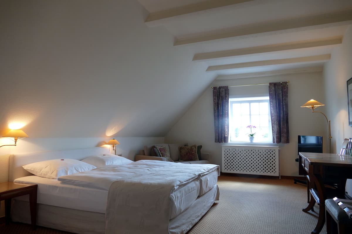 A bedroom with a large bed, bedside lamps, a window with curtains, a radiator, a desk, and a television.