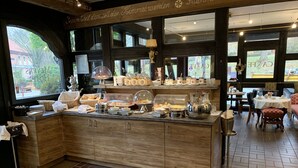 A breakfast buffet area with a wooden counter, various food items, and a view of the outdoors through large windows. - Meyers Hotel (Seevetal)