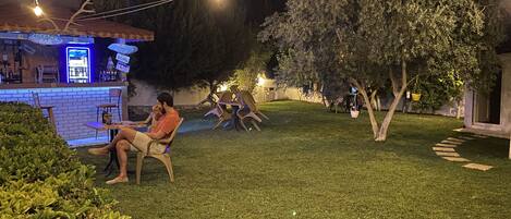 A well-lit outdoor seating area with a bar, chairs, and a grassy lawn.