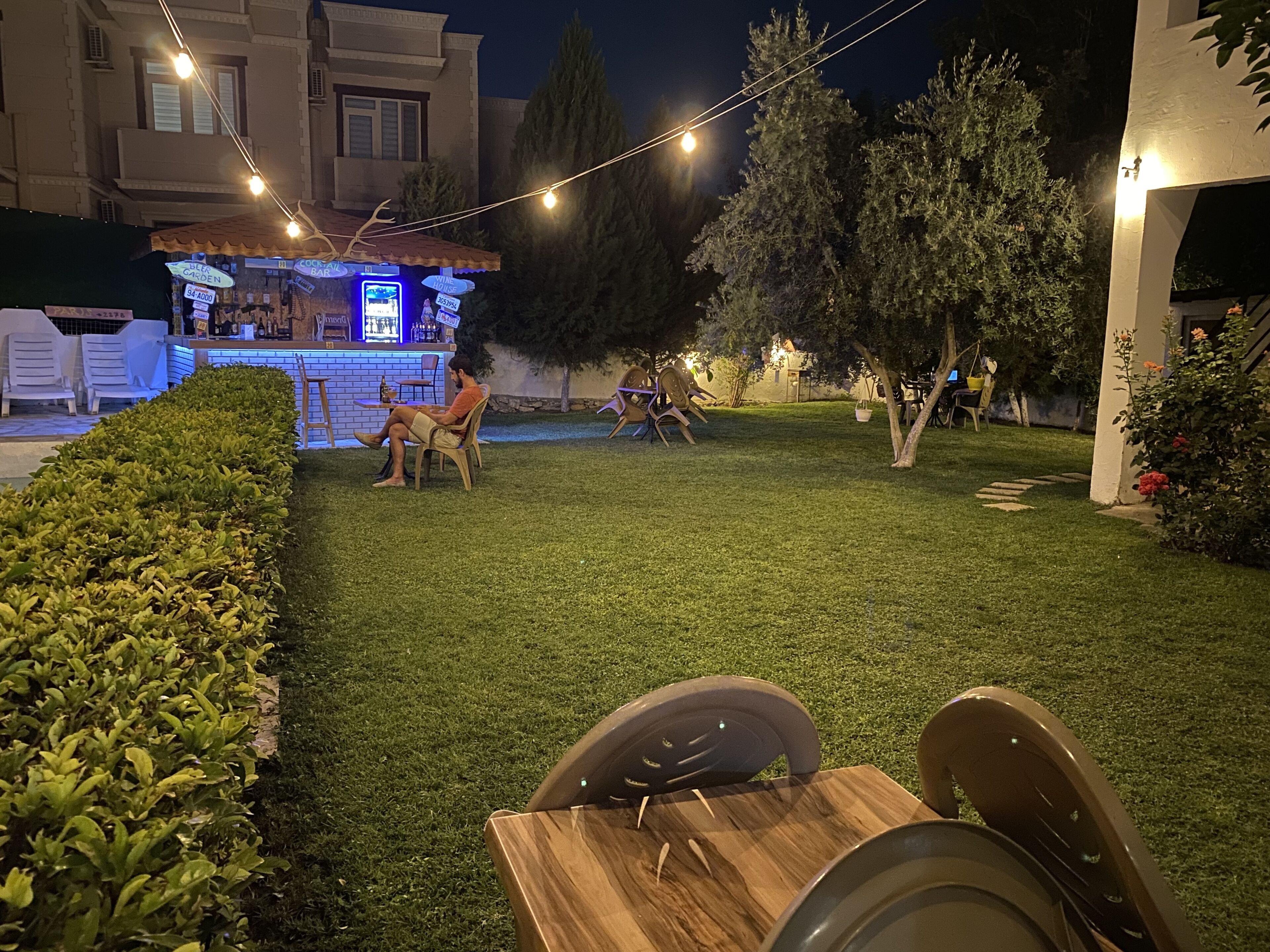 Un'area salotto all'aperto con tavoli e sedie, una zona bar con insegne al neon e un prato ben curato.