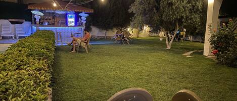 Un'area salotto all'aperto con tavoli e sedie, una zona bar con insegne al neon e un prato ben curato.