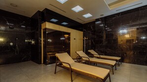 A modern spa room with black marble walls, a wooden cabinet, and a glass door leading to a sauna area. There are four lounge chairs with beige cushions. - Golden Palace Batumi Hotel & Casino (Batumi)