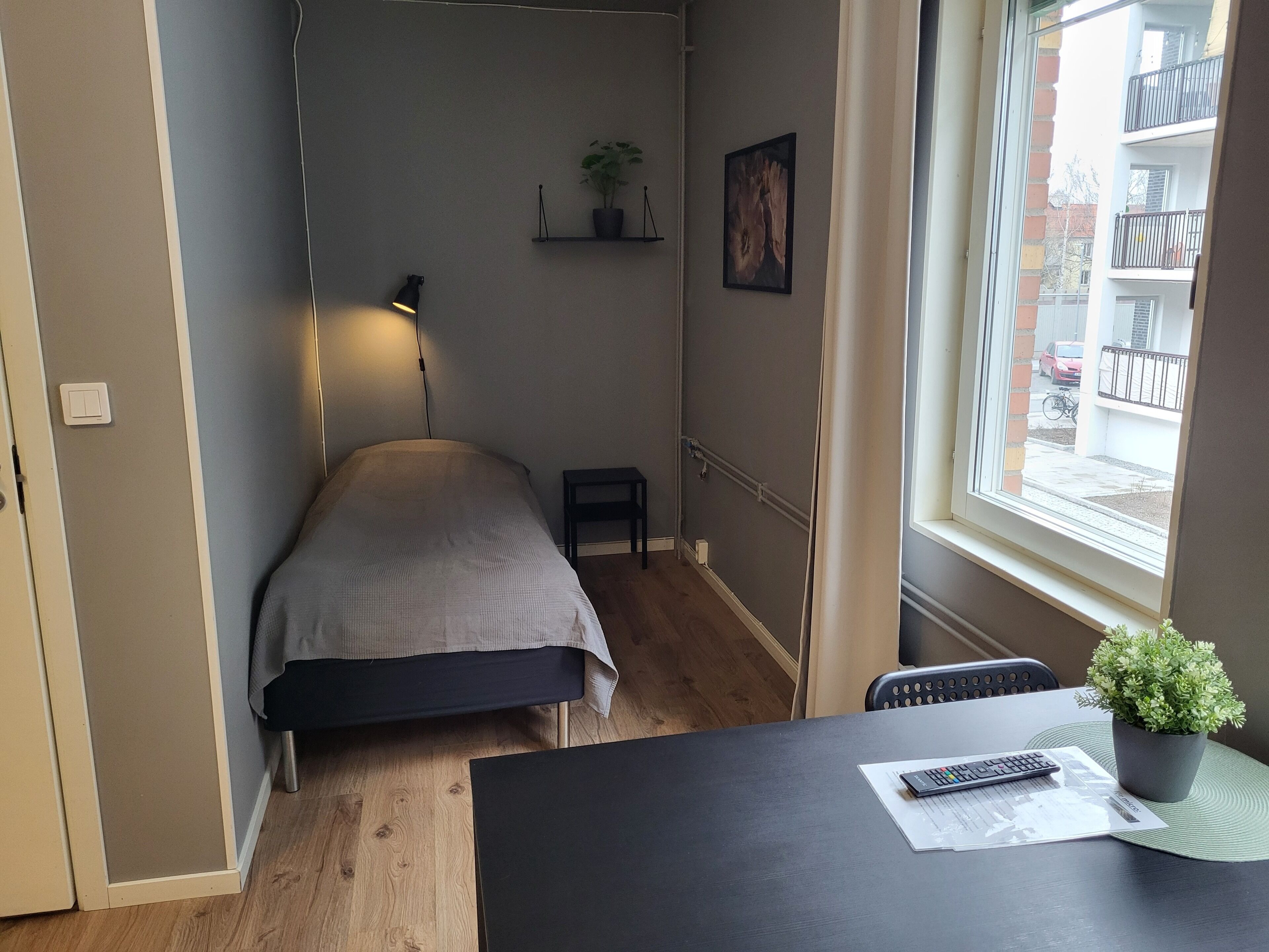 Una habitación pequeña y funcional con cama, escritorio con planta, lámpara y ventana con vista a edificios.
