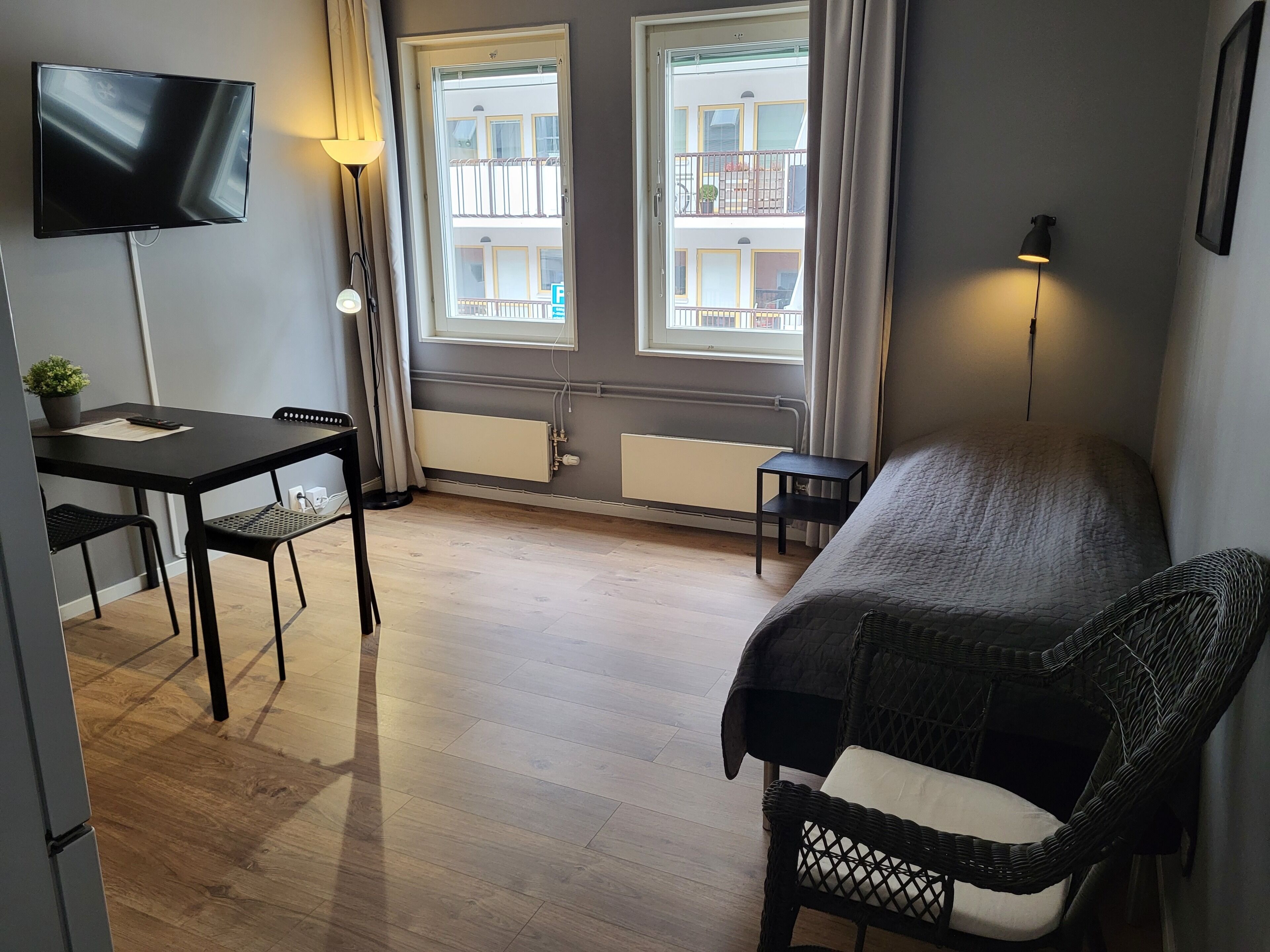 A room with a bed, a TV, a table with chairs, a small table with a lamp, and a window with a view of buildings.