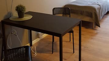 A room with a bed, a table with a potted plant, two chairs, a television mounted on the wall, and a window with curtains.