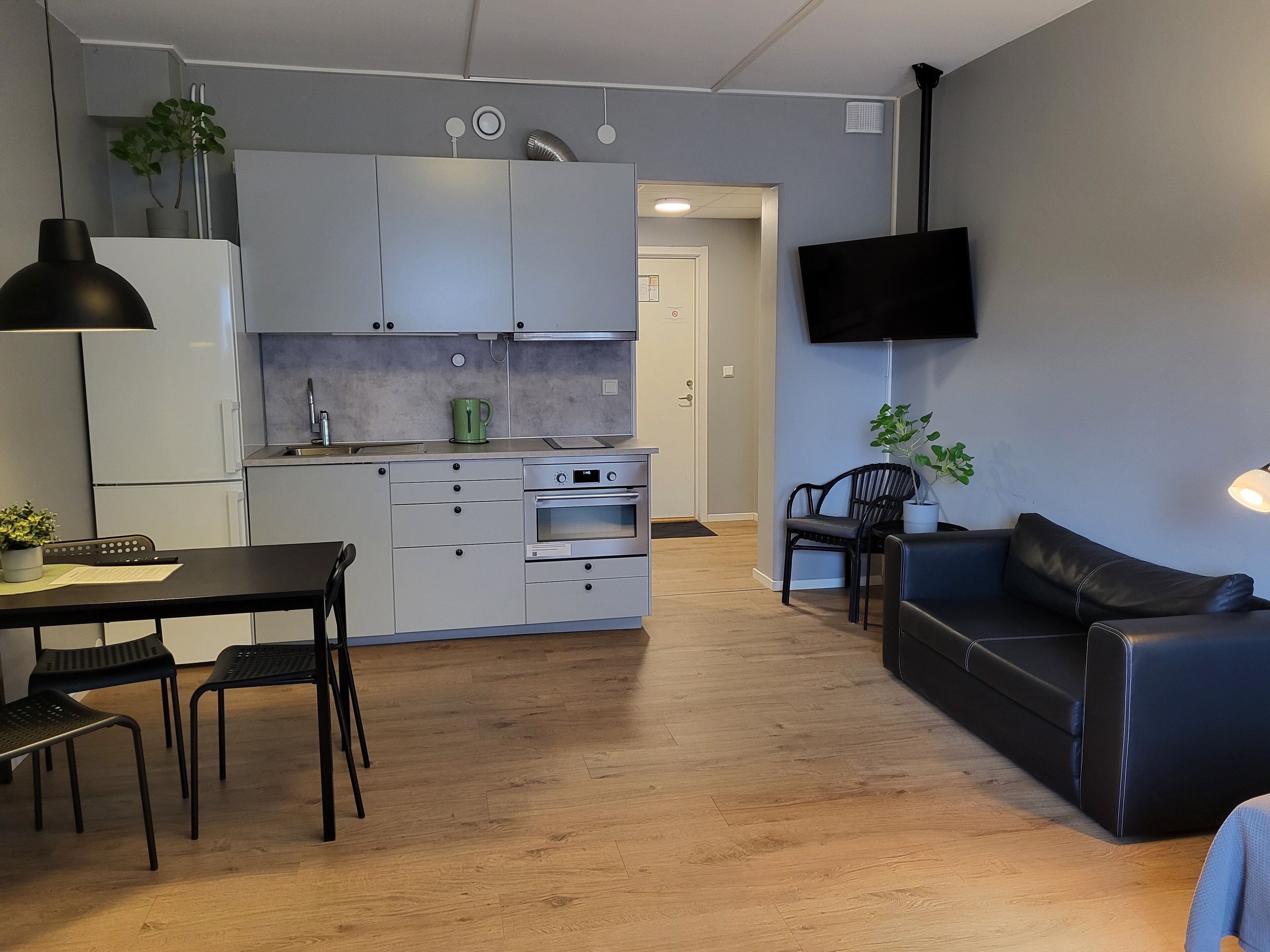 Una cocina moderna con armarios blancos, una mesa de comedor con sillas, un sofá negro y un televisor montado en la pared.