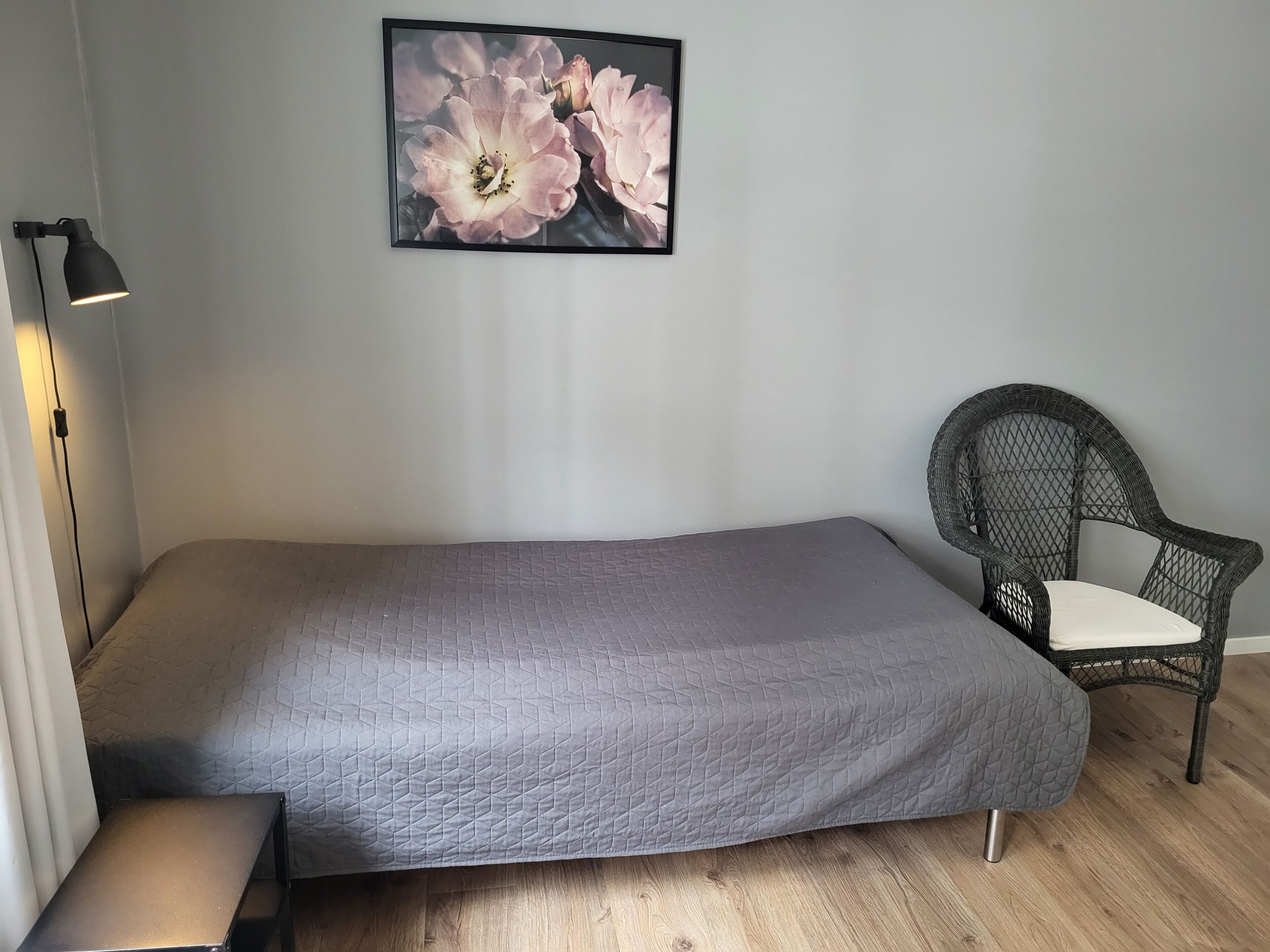 Una cama individual con colcha gris, una silla de mimbre con cojín blanco, una lámpara de pie negra y una obra de arte floral enmarcada en la pared.