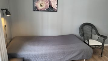 Una cama individual con colcha gris, una silla de mimbre con cojín blanco, una lámpara de pie negra y una obra de arte floral enmarcada en la pared.