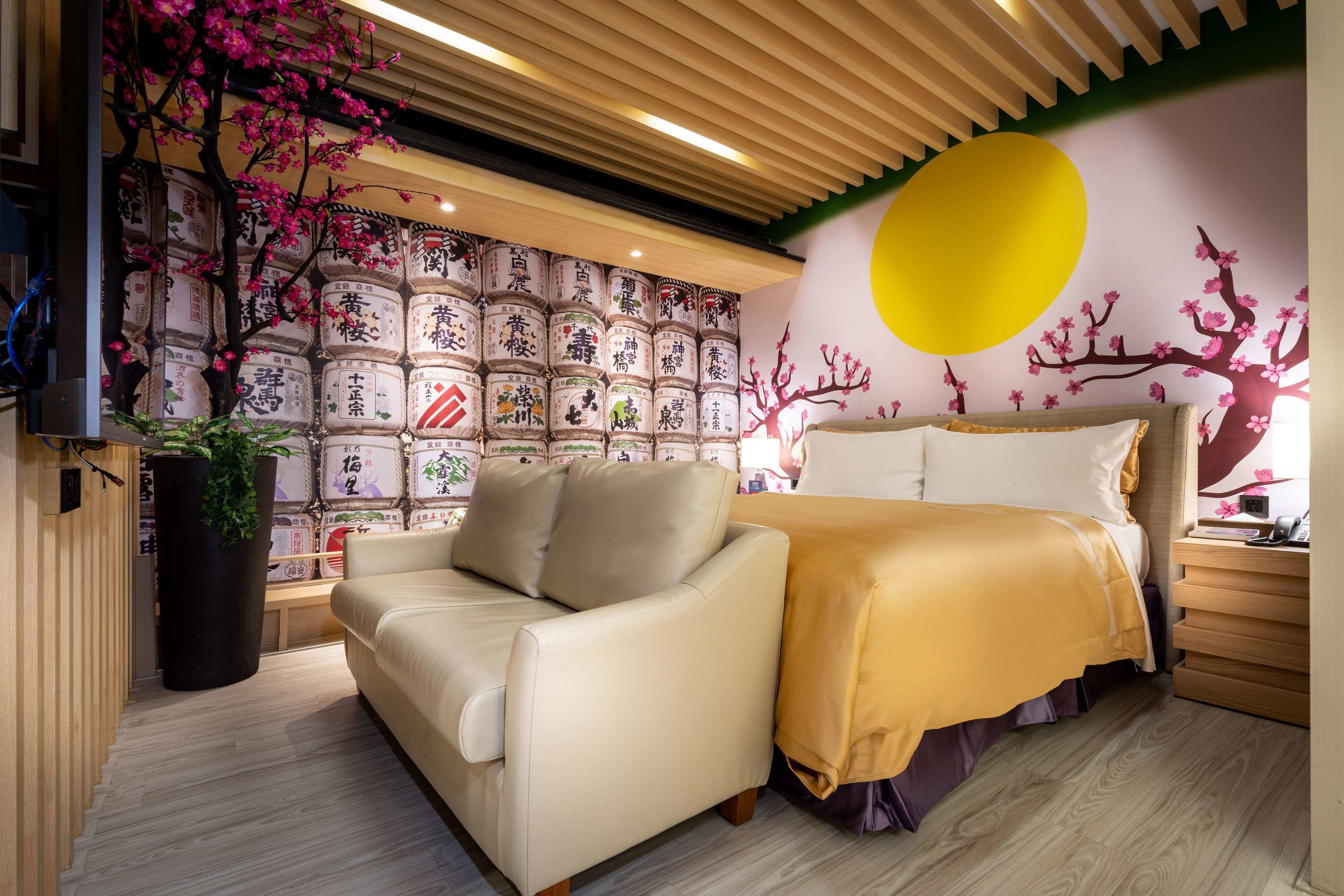 A modern hotel room with a sofa, a bed with a yellow bedspread, a large wall mural with traditional Japanese elements, and a wooden ceiling.