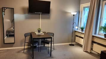 Et kompakt værelse med et fladskærms TV på væggen, et spisebord med to stole, en gulvlampe og et vindue med gardiner.