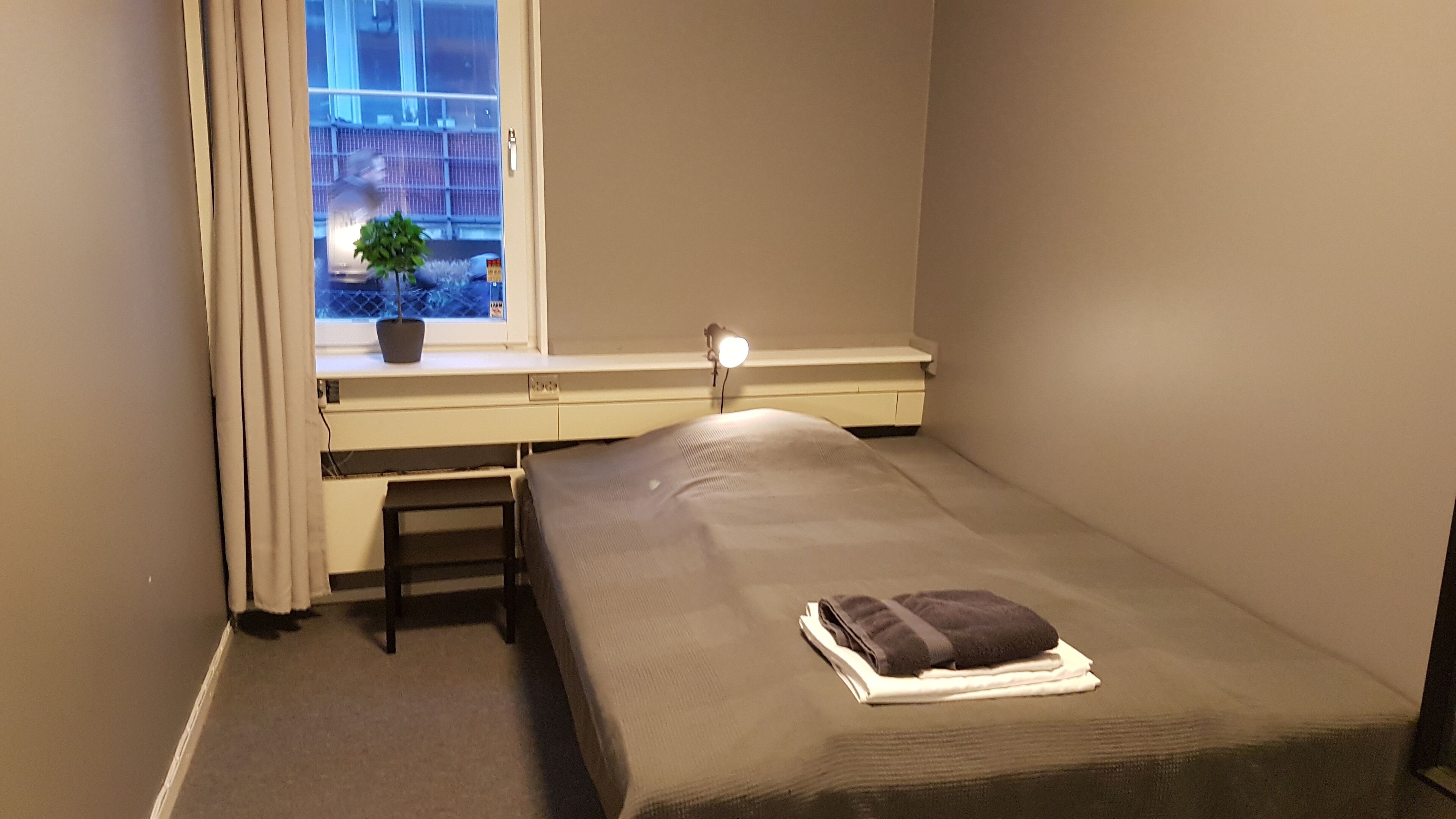 A small, single-bed room with a desk and chair, a window with a view of a building, and a bedside lamp.