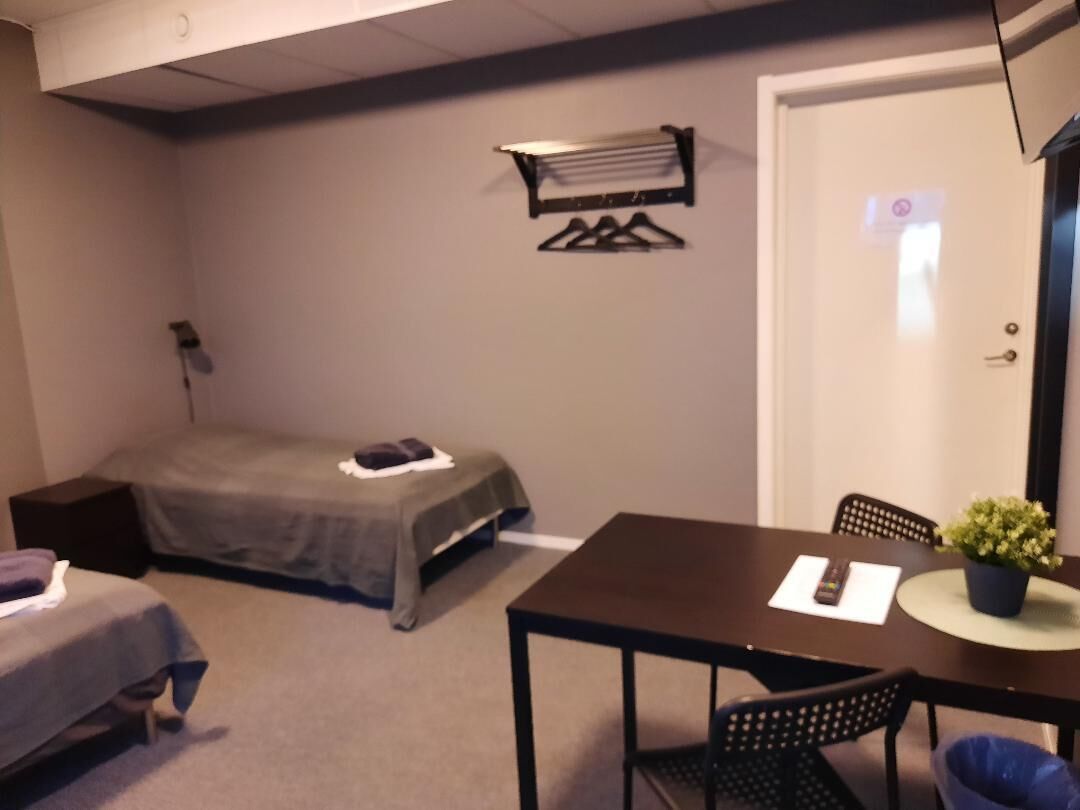 Una habitación pequeña y ordenada con cama, una mesa con sillas, una planta en maceta y un perchero.