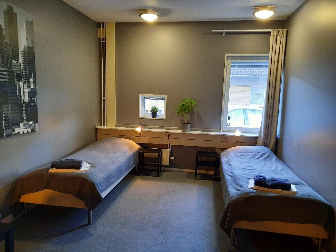 A small, compact room with two single beds, a desk with a chair, a window with curtains, and a small potted plant.