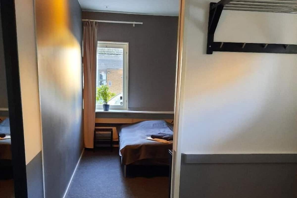 Una habitación pequeña con cama, una mesita, ventana con cortinas y un radiador.