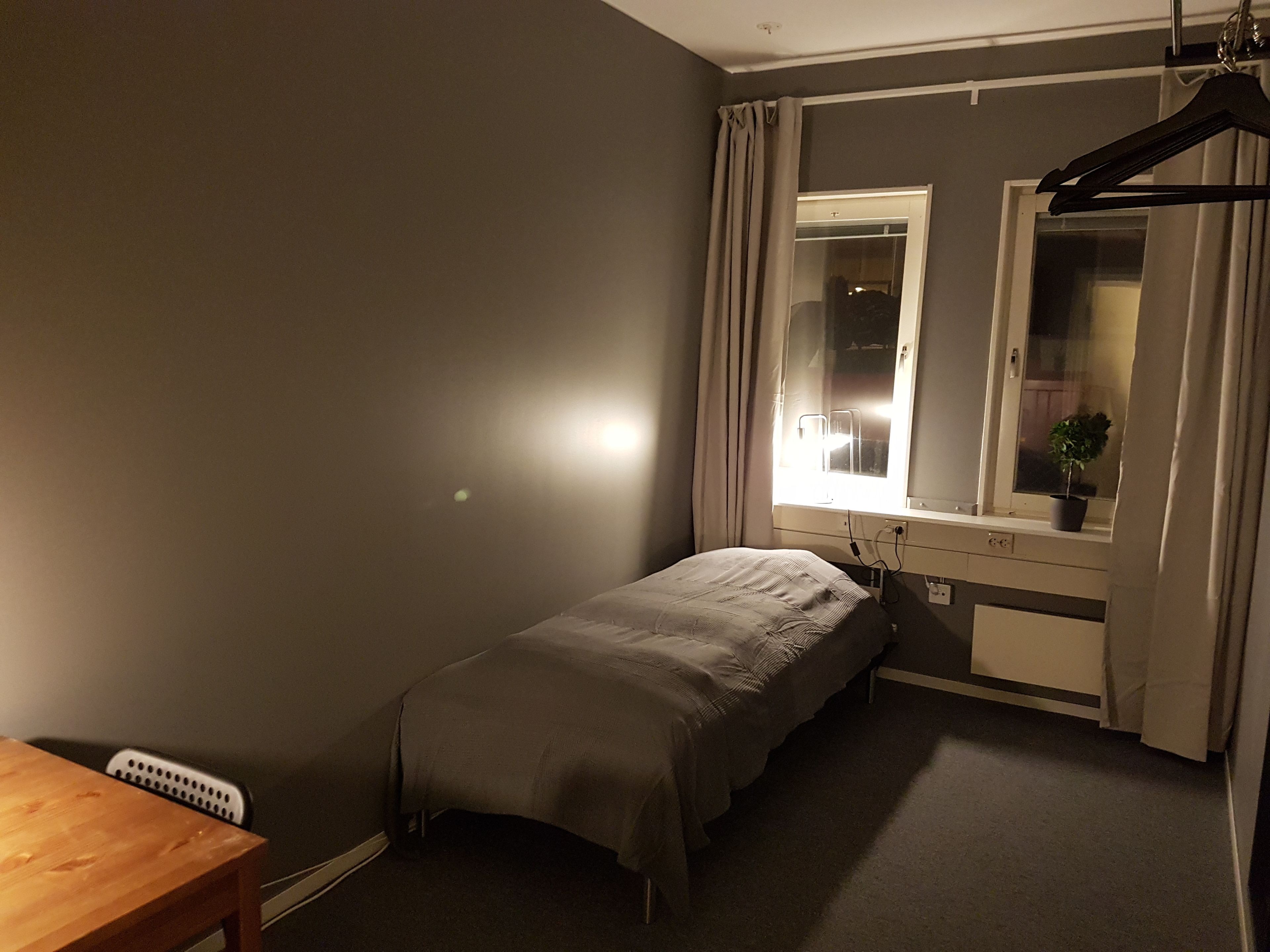 A small, dimly lit bedroom with a single bed, a window with curtains, a radiator, and a wooden table.