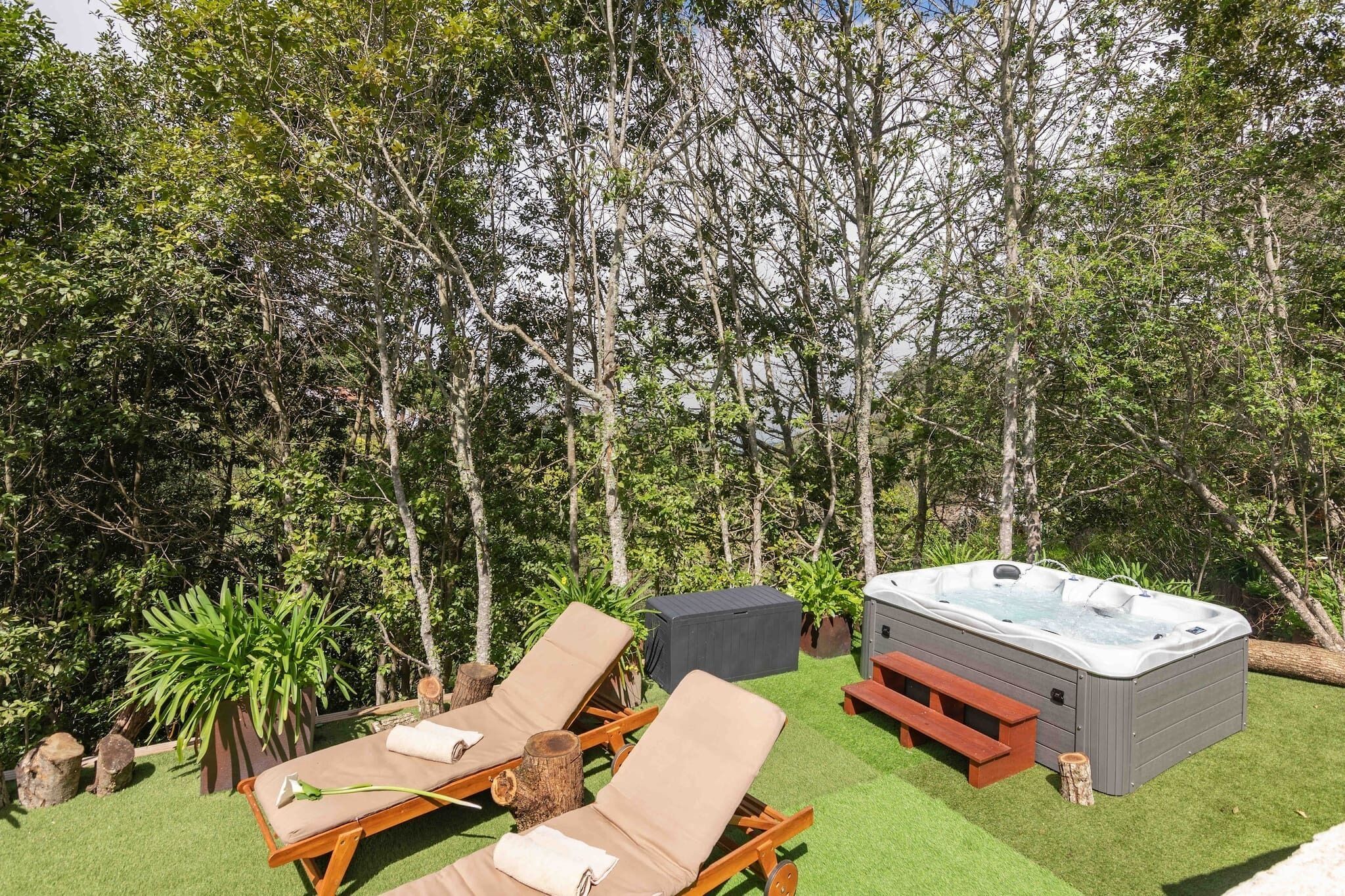 A hot tub surrounded by lounge chairs and a picnic table on a grassy lawn with trees in the background.