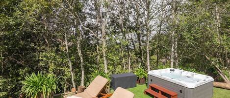 A hot tub surrounded by lounge chairs and a picnic table on a grassy lawn with trees in the background.