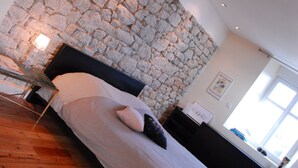 A bedroom with a stone wall, a bed with pillows, a bedside table, and a window. - Villa Tourelle (Dinan)