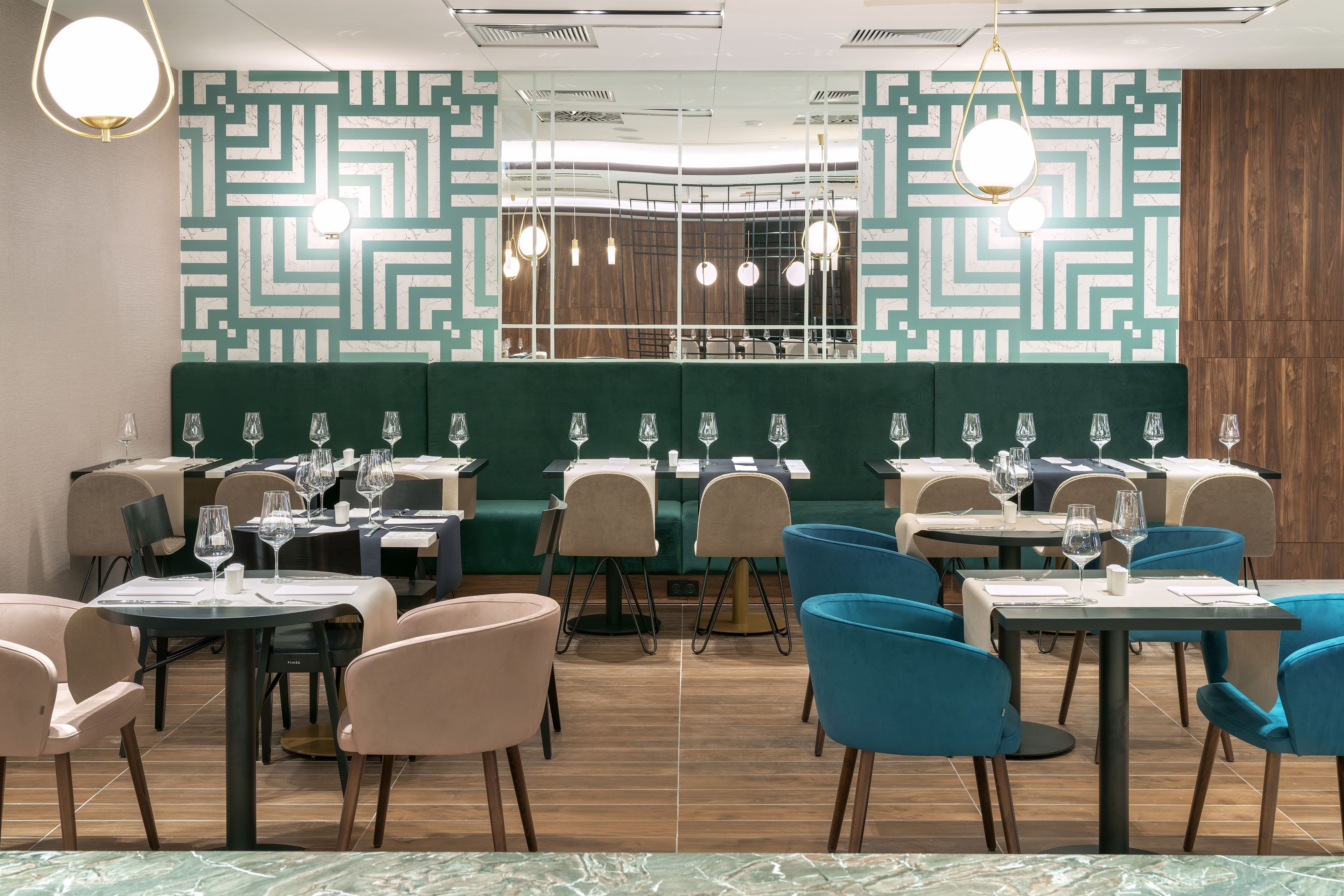 Un restaurant aux murs ornés de motifs verts, au plancher en bois et aux chaises bleues et beiges. Les tables sont dressées avec des verres à vin et des couverts.