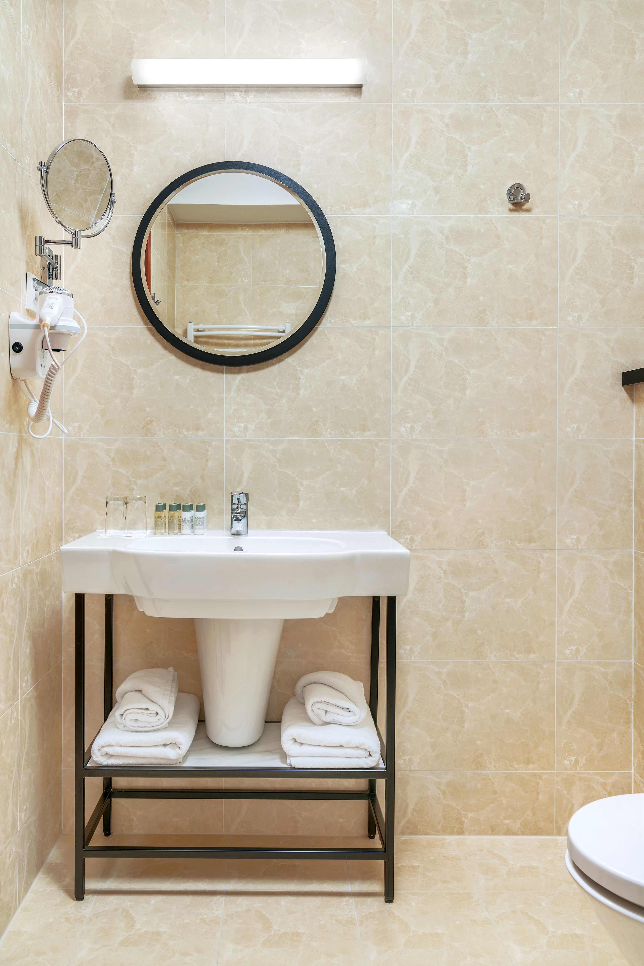 Un baño con lavamanos blanco, un espejo redondo y un perchero con toallas dobladas.