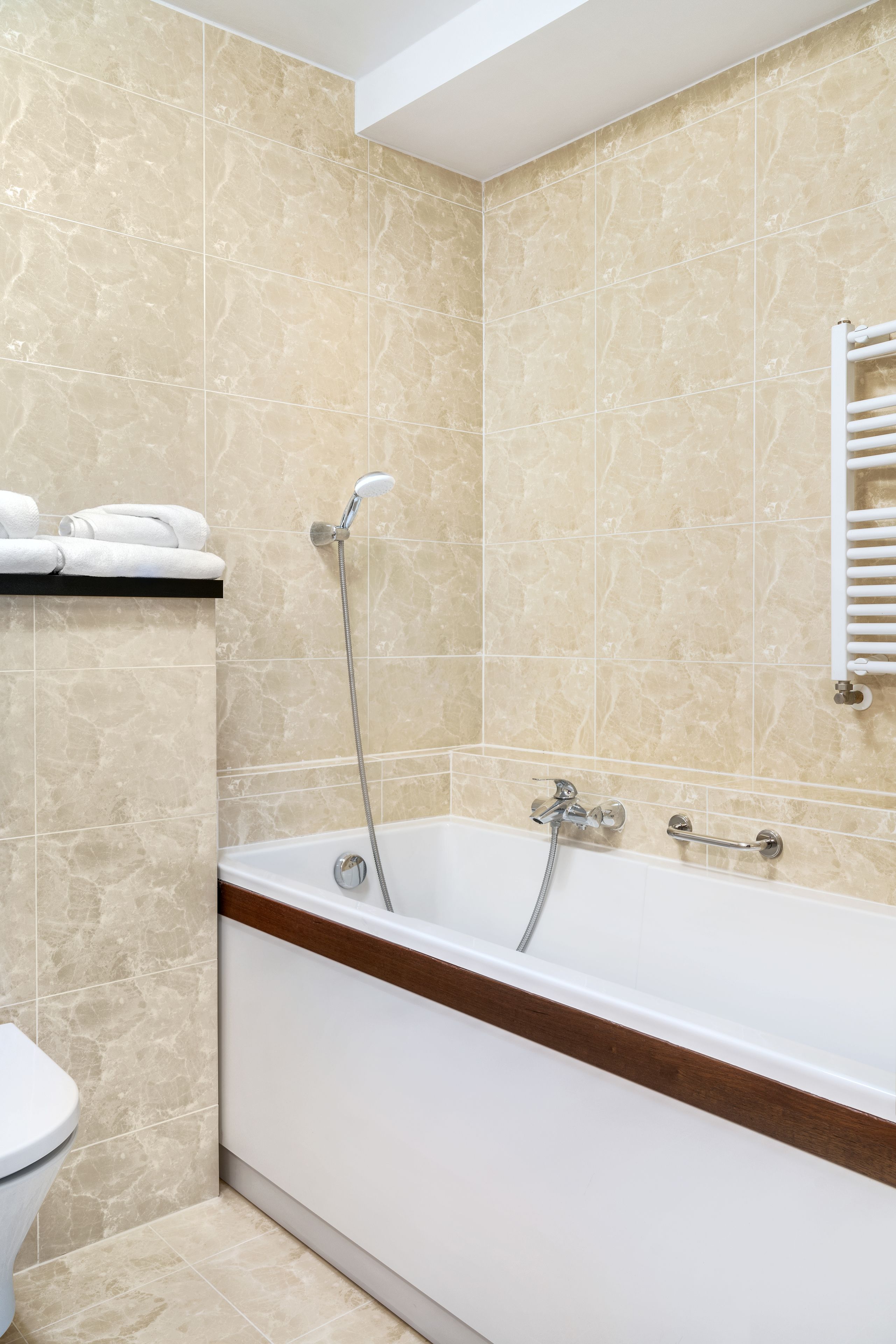 A bathroom with beige marble tiles, a white bathtub with a wooden edge, a white towel rack, and a white toilet.