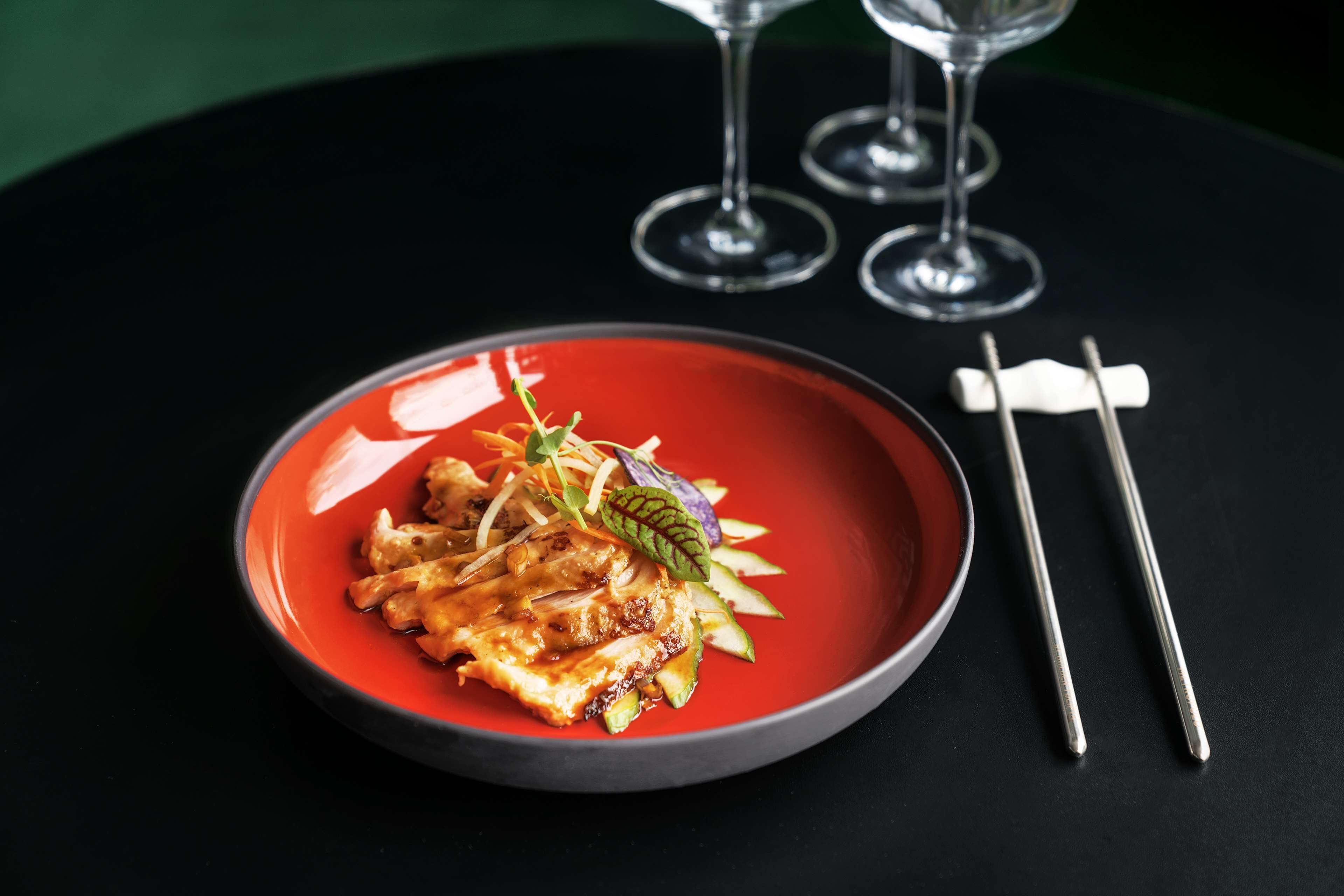 A red plate with food on a black table, accompanied by chopsticks and wine glasses.
