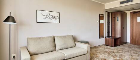 A neutral-toned sofa with a side table, a floor lamp, a framed artwork, and a wooden cabinet in a room with a patterned carpet.