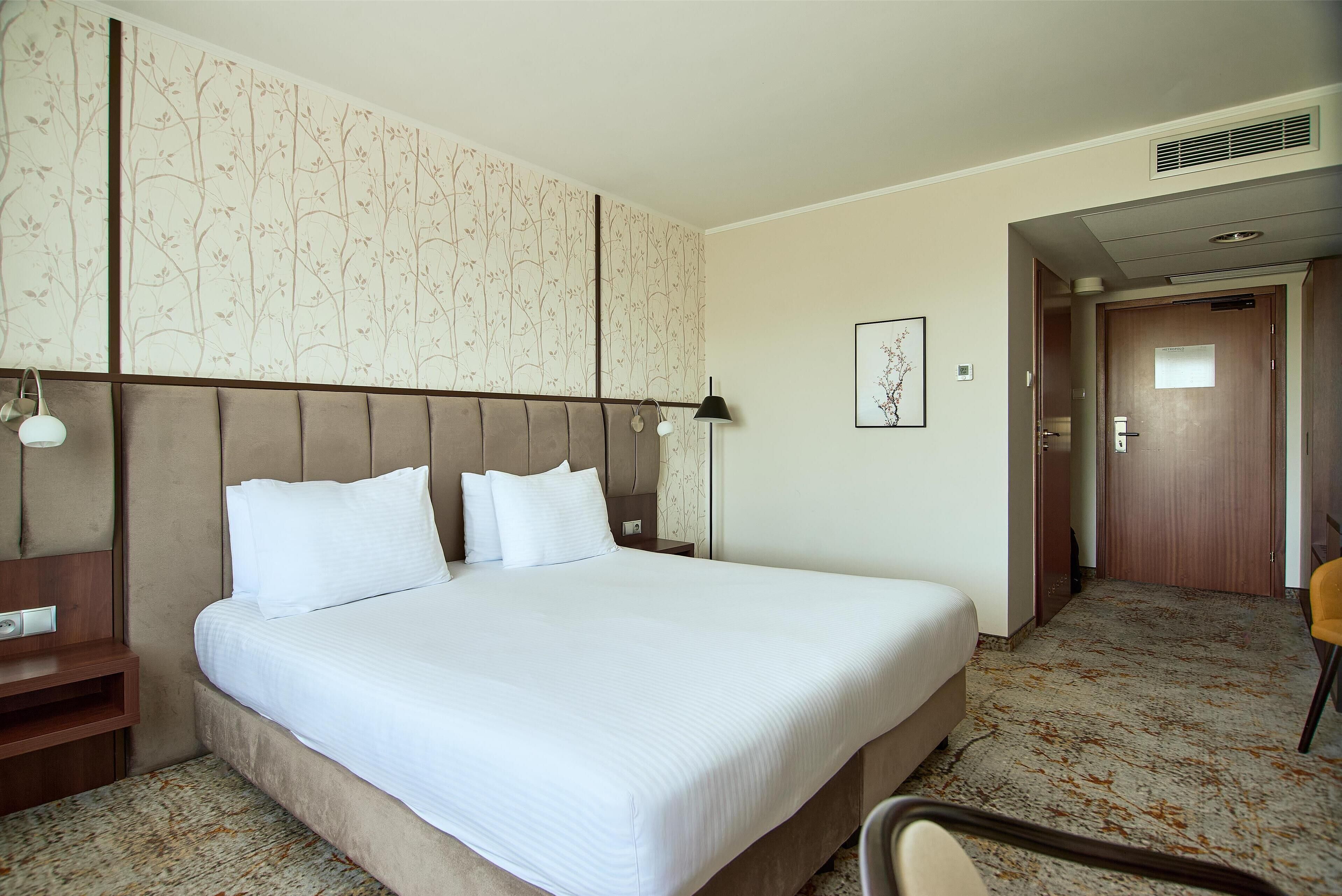 Habitación de hotel con una cama grande, una mesita de noche, una lámpara, un cuadro en la pared y una puerta que da a otra habitación.