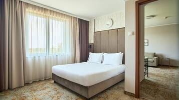 Habitación de hotel con una cama grande, vista al exterior a través de cortinas translúcidas, cabecera con un reloj y piso alfombrado.