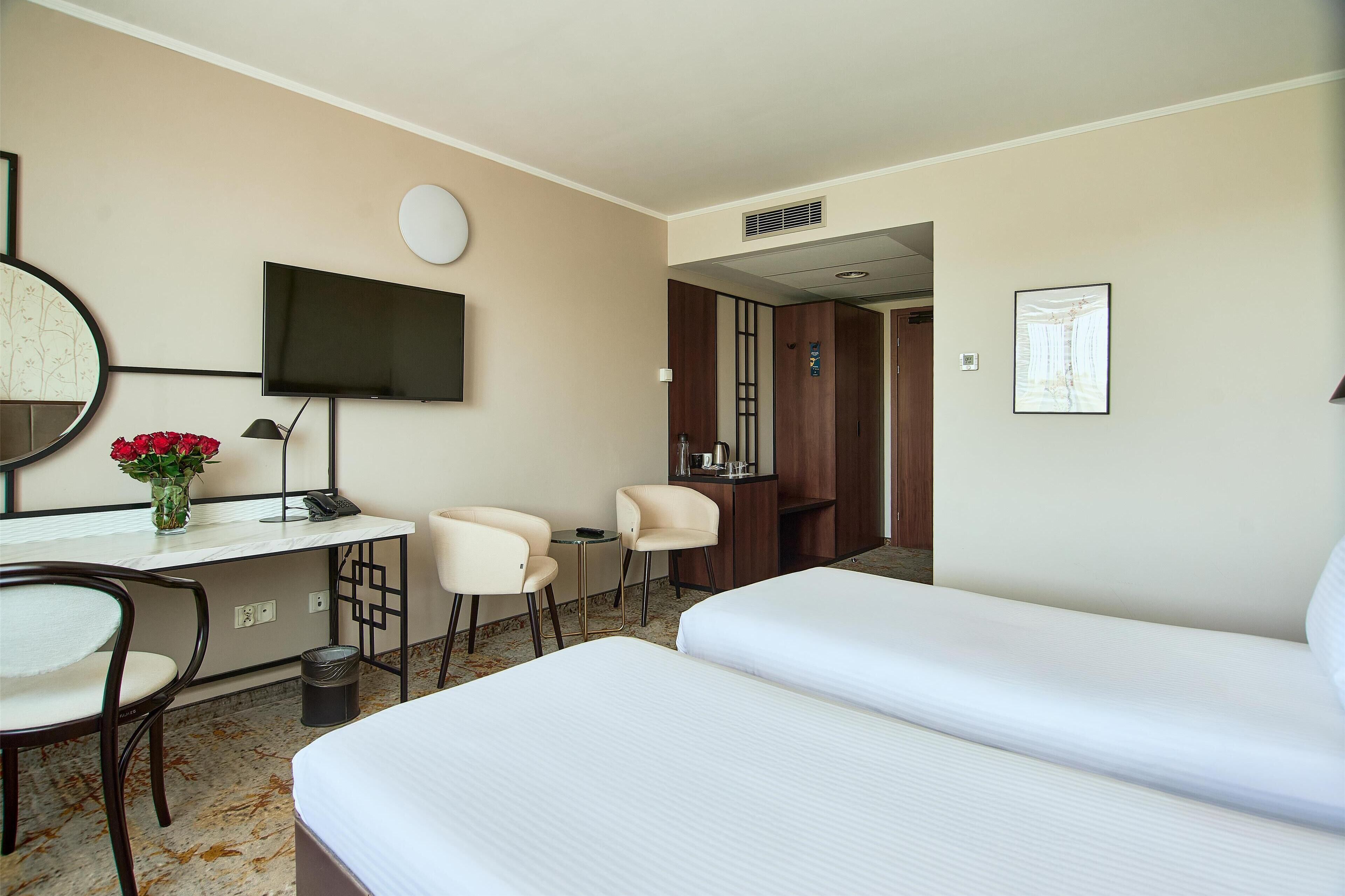 A hotel room with two beds, a desk with a chair, a TV, and a small table with a vase of flowers.