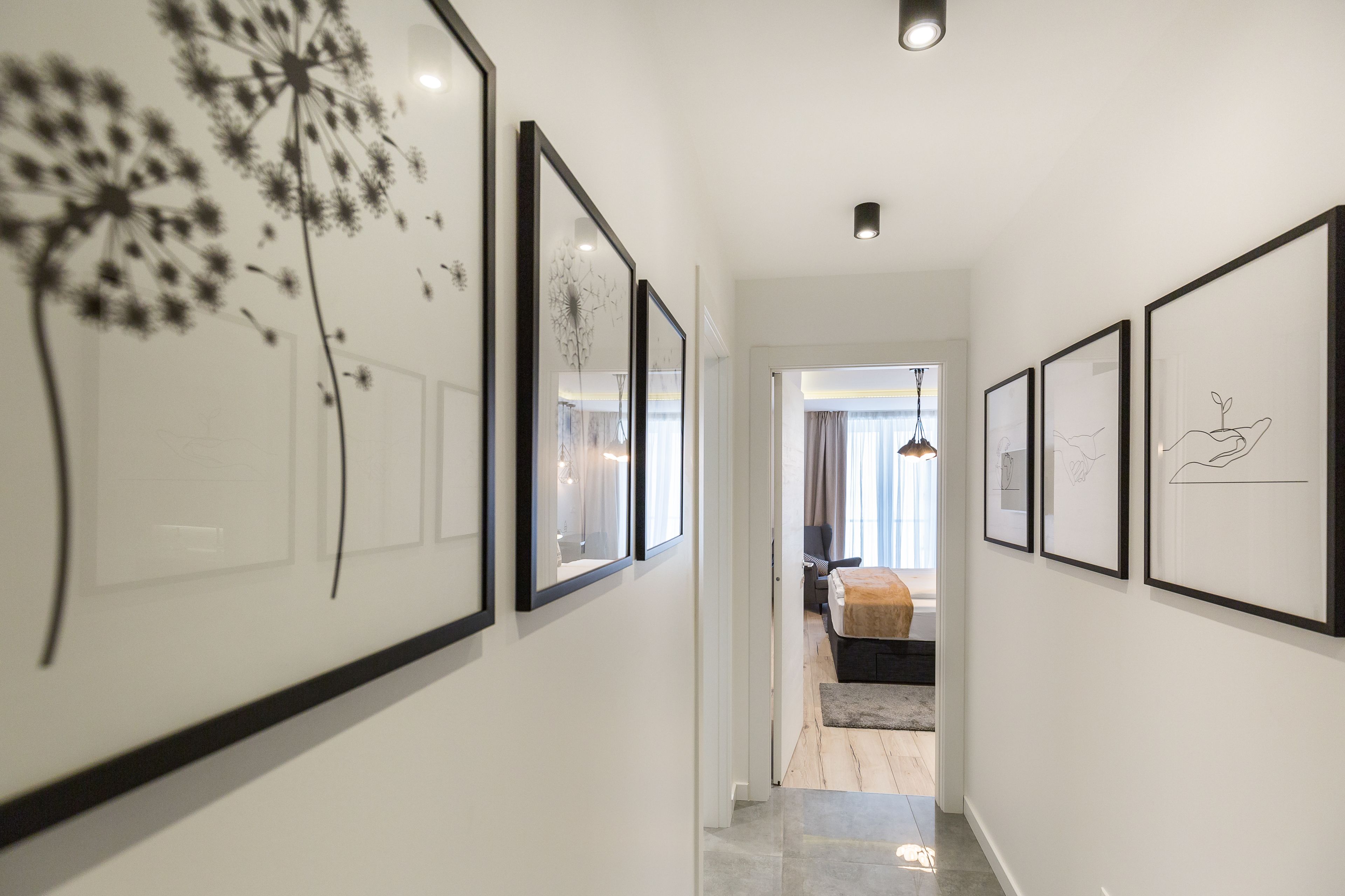 A hallway with framed artwork on the walls, leading to a bedroom with a bed and a window.