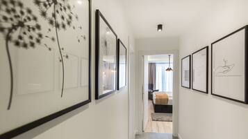 Un couloir orné de tableaux encadrés le long des murs, qui mène à une chambre à coucher avec un lit et une fenêtre.