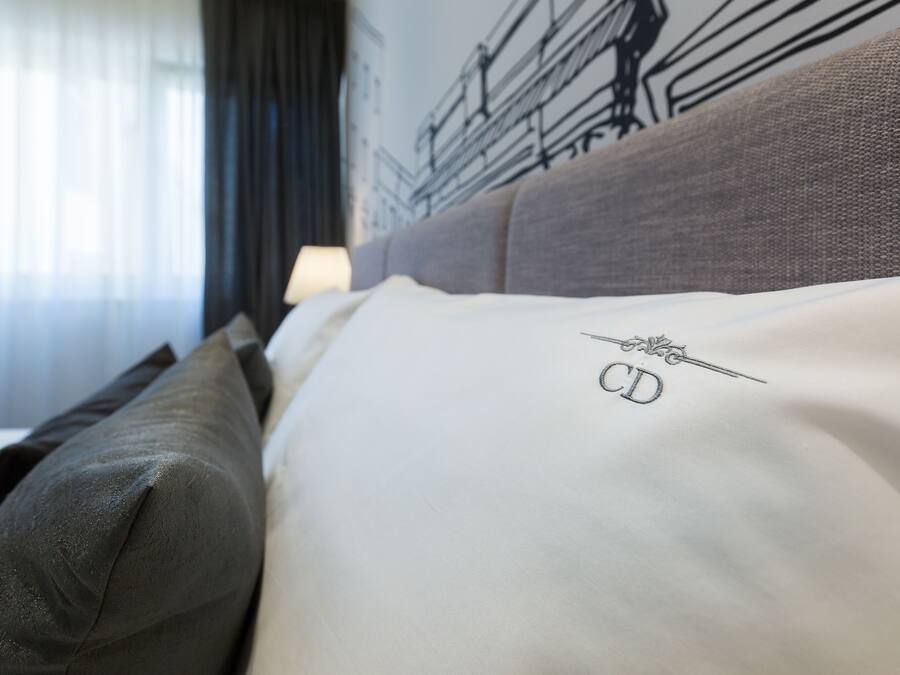Habitació d'hotel amb un llit, amb una capçalera gris i un coixí blanc amb text brodat.