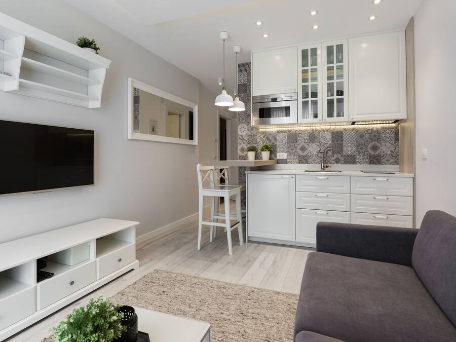Een moderne woonkamer met een flatscreen televisie, een witte entertainmentkast, een grijze bank en een keuken met witte kasten en een decoratieve spatwand.