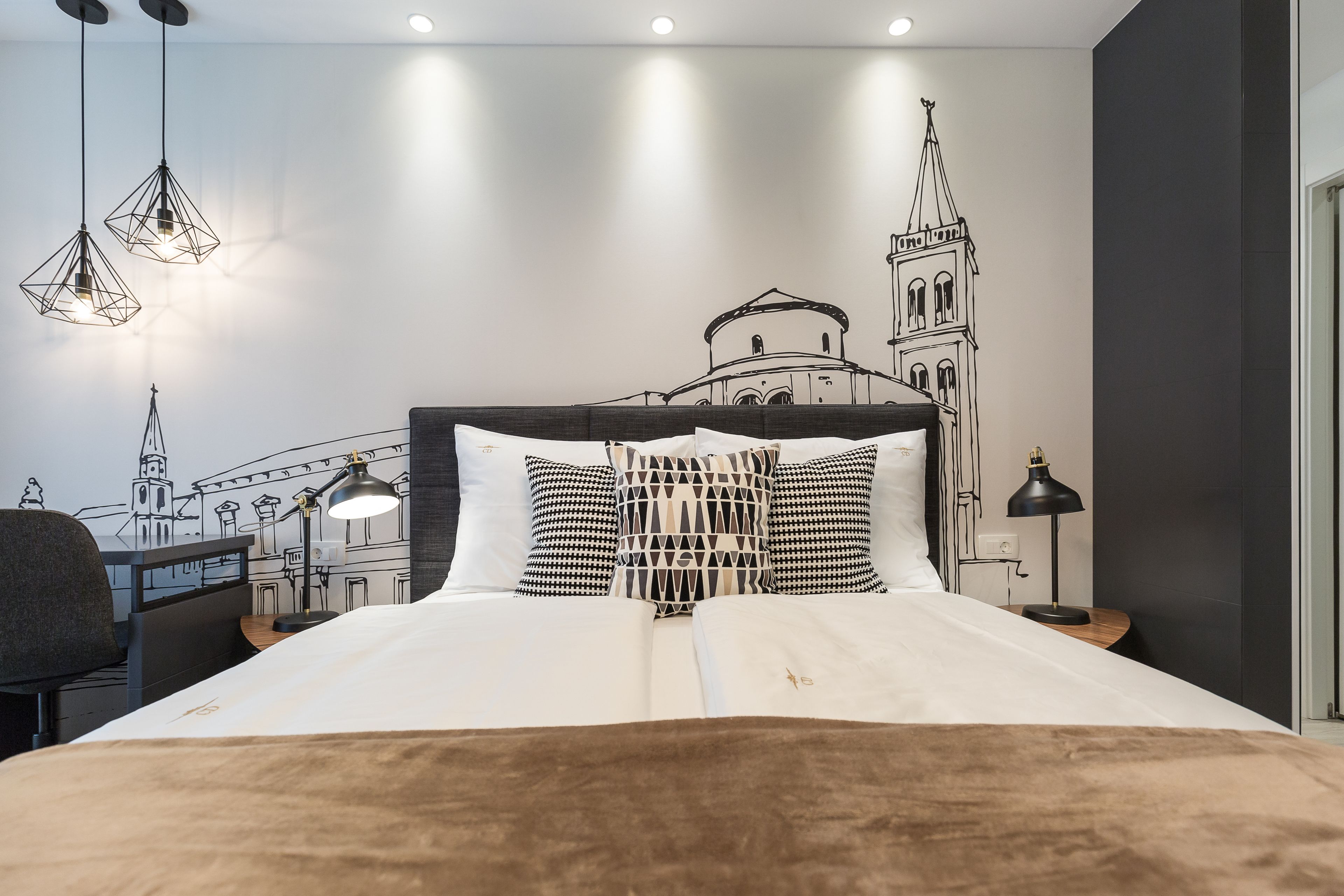 Ein Hotelzimmer mit Bett, Schreibtisch und Stuhl. An der Wand befindet sich ein Wandbild eines Gebäudes, und über dem Bett hängen drei Pendelleuchten.