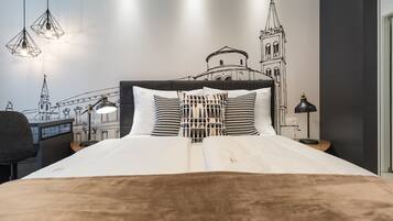 Ein Hotelzimmer mit Bett, Schreibtisch und Stuhl. An der Wand befindet sich ein Wandbild eines Gebäudes, und über dem Bett hängen drei Pendelleuchten.