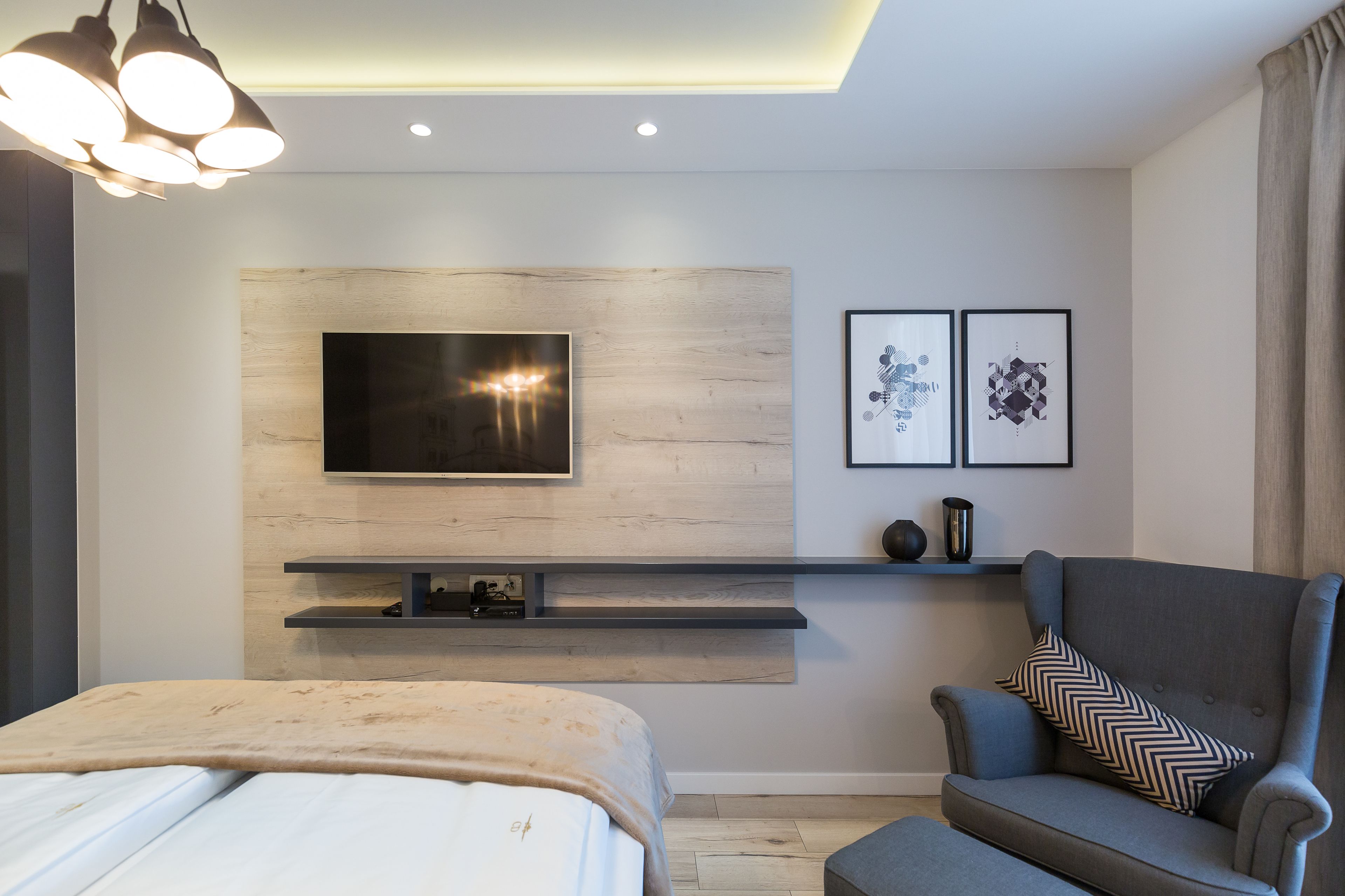 A modern bedroom with a bed, a grey armchair, a TV mounted on a wooden panel, and a shelf with decorative items.